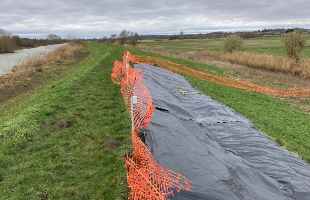A picture showing the rivers embankment showing Tilt sensors installed at the top of an embankment, above an area prone to landslip which is covered with a temporary plastic cover, with the area of the emergency earthworks monitoring fenced off to prevent access to the public.