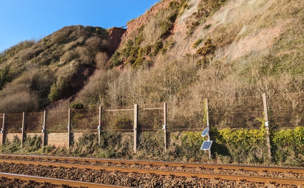 An image taken from the track, showing the location of the emergency cliff monitoring. A solar panel can be seen trackside while a fence can be seen hanging above the area of the landslip at the top of the cliff